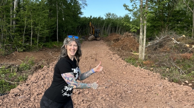 alicia is standing in front of a roughed in driveway on wooded land smiling and giving the thumbs up