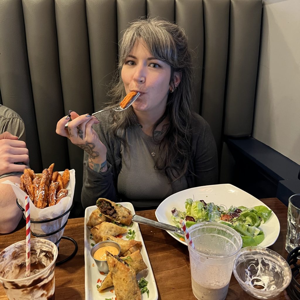 Alicia is sitting at a booth putting a sweet potato fry to her mouth with a fork and is surrounded by plates of food