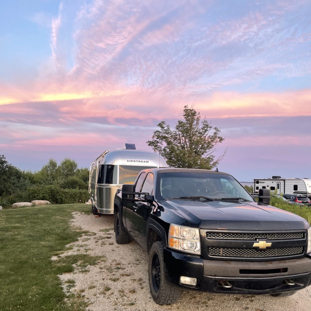 A beautiful pastel colored sunset sky sits behind the airstream trailer with a black chevrolet silverado in front of it on a grassy field