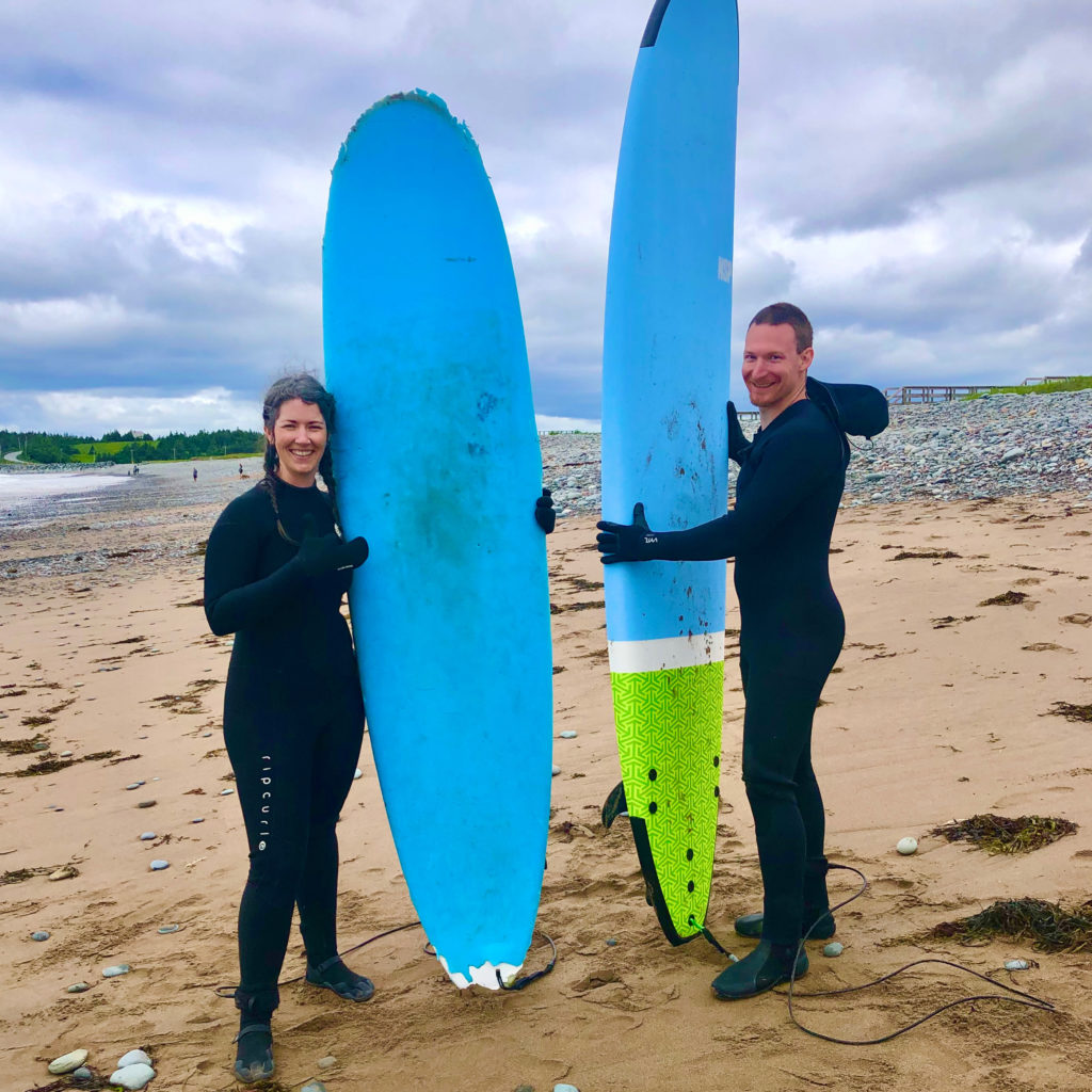 A man and a woman in black wetsuits standing on Lawrencetown Beach holding bright blue surf boards.